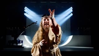 Eine bärtige Gestalt in einem beigen Gewand steht auf der Bühne und zeigt in die Kamera - Annina Machaz als Jesus in Florentina Holzingers Opernperformance "Sancta"