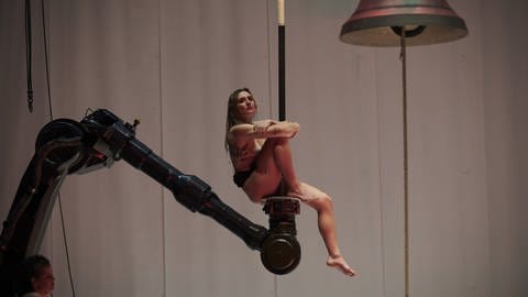 Eine nackte Frau Sitzt auf einem Kran, neben ihr eine Glocke - Szene aus Florentina Holzingers Opernperformance "Sancta"