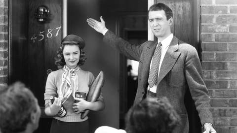 Eine Aufnahme aus einem Film in schwarz-weiß mit James Stewart und Donna Reed vor einem Hauseingang. Stewart gestikuliert im Anzug und zeigt auf eine Austür.