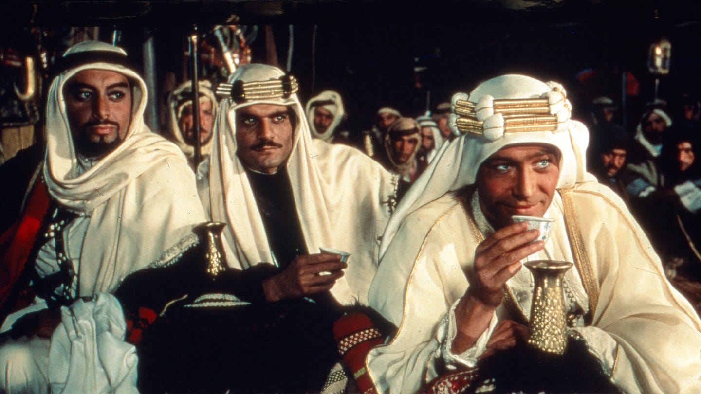 Moviestill von Lawrence von Arabien. Lawrence (Peter O'Toole) sitzt im arabischen Outfit vorne im Bild und trinkt Tee.