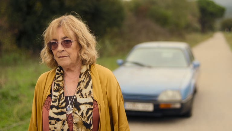 Eine Frau mit Sonnenbrille ist zu sehen, im Hintergrund ein blaues Auto (Filmstill zur Serie "Land of Women" auf Apple TV+)