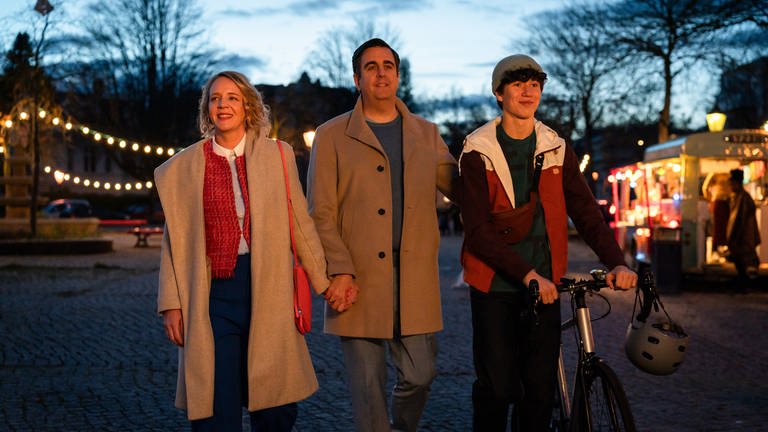 "Alles gelogen": Am Abend: Vera (Katrin Wichmann), Hajo (Bastian Pastewka) und Marvin Siewers (Arthur Gropp) laufen lächelnd nebeneinander über einen mit Lichterketten geschmückten Platz. Marvin schieb ein Fahrrad. Im Hintergrund steht ein beleuchteter Foodtruck.
