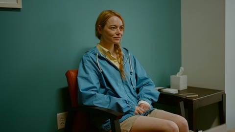 Emma Stone sitzt auf einem Stuhl und lächelt im Film KINDS OF KINDNESS