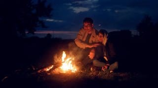 Geflüchtete (Mann und Frau) sitzen an Lagerfeuer, Filmszene aus "Echoes from Borderland"