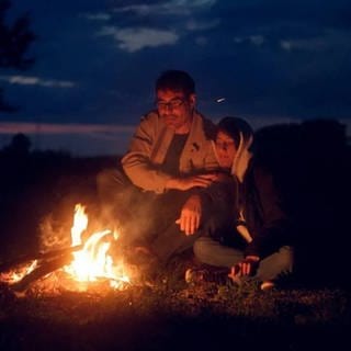 Geflüchtete (Mann und Frau) sitzen an Lagerfeuer, Filmszene aus "Echoes from Borderland"