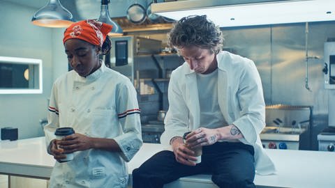 Sydney (Ayo Edebiri) und Carmy (Jeremy Allen White) sitzen auf der Kücheninsel in der Küche des Restaurants. Sie trinken Kaffee und schauen betreten auf ihre Becher.