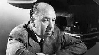 Regisseur Alfred  Hitchcock (1899-1980) sitzt, die Arme auf die Rückenlehne seines Regiestuhls gelehnt, vor einer Filmkamera.