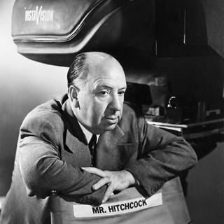 Regisseur Alfred  Hitchcock (1899-1980) sitzt, die Arme auf die Rückenlehne seines Regiestuhls gelehnt, vor einer Filmkamera.