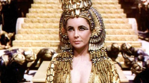 „Cleopatra“ (1963): Kleopatra (Elizabeth Taylor) in ihrem Pharaoninnenornat, im Hintergrund eine pyramidenartige Prunkkutsche für den Triumphzug der Monarchin.