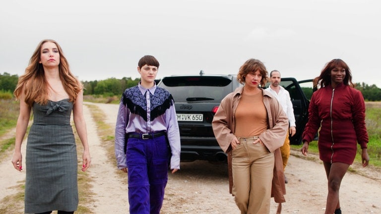 Filmstill: Vier Frauen laufen selbstbewusst eine Feldstraße entlang. Im Hintergrund sind ein Auto und ein Mann zu sehen. 