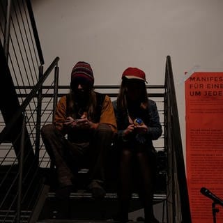 zwei Menschen sitzen auf einer Treppe, daneben ein großes rotes Plakat