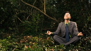 Young businessman sits in a lotus position meditating in the woods with closed eyes and a smile.