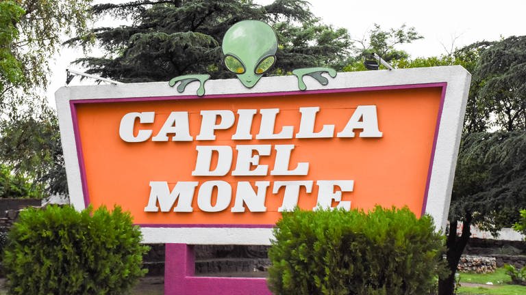 Ortsschild von Capilla del Monte. Auf dem Ortsschild sitzt die Figur eines Außerirdischen.