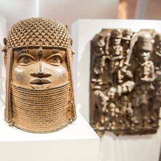 In einer Vitrine sind mehrere sogenannte „Benin-Bronzen“ auf weißem Untergrund ausgestellt. Im Vordergrund ist eine goldglänzende Büste eines geschmückten Kopfes. Im Hintergrund sind zwei Bronze-Relieftafeln an weißen Stellwänden aufgehängt.