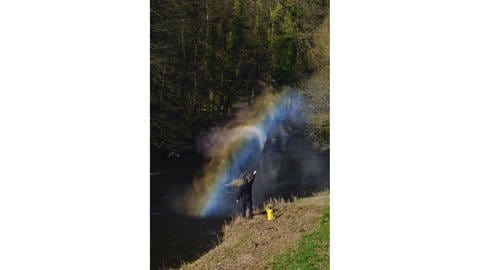 Eine am Ufer eines Flusses stehende Person, sprüht einen feinen Wasserdunst in den Himmel und zaubert so einen Regenbogen