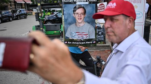 Mann fotografiert sich und eine demonstrierende Person, die ein Bild des Trump-Attentäters Thomas Matthew Crooks hochhält, auf dem dieser als amerikanischer Held bezeichnet wird.