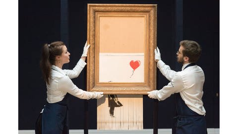 Das geschredderte Banksy-Bild "Love is in the Bin" wird vor der Auktion von zwei Menschen mit Handschuhen an eine Wand gehängt.