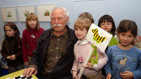 Der Künstler Janosch mit mehreren Kindern, ein Mädchen hält ein Schild mit einer Tigerente in die Höhe