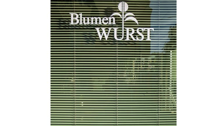 Vor einer grünen Jalousie-Lamellen eine stilisierte Pflanze mit der Aufschrift „Blumen Wurst“ 