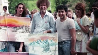 Bob Ross umgeben von Fans. Sie halten selbstgemalte Bilder in den Händen. 