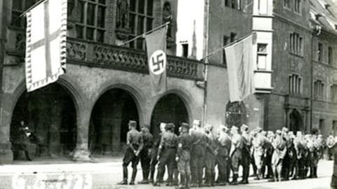 Männer stehen in Reih und Glied vor einem Gebäude, von dessen Balkon drei große Fahnen hängen