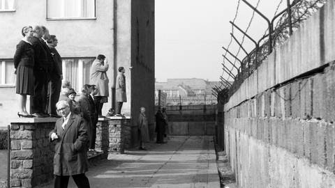 Eine Reisegruppe schaut von einer Vorgartenmauer über die Mauer hinweg vom Westteil in den Ostteil von Berlin, Deutschland 1962: Schwarz-weiß Bild, links stehen die Menschen, Männer und Frauen auf einem kleinen Mäuerchen, rechts befindet sich in mehreren Metern Abstand die Mauer, die oben mit Stacheldraht gesichert ist.