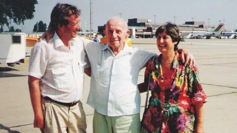 Israel Shiloni in der Mitte des Bildes. Er legt die Arme um einen Mann links im Bild und eine Frau rechts im Bild. 