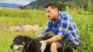 Christian Schindler sitzt auf einem großen Stein auf einer Wiese. Vor sich ein schwarzer Hund, den er streichelt.