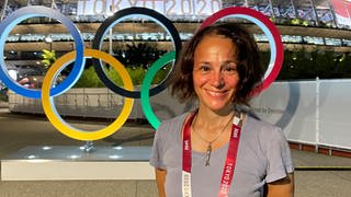 Kathrin Erdmann vor dem Nationalstation in Tokio. Hinter ihr die Olympischen Ringe in den Farben blau, gelb, scharz, grün und rot. Auf einem Banner am Stadion steht "Tokio 2020" geschrieben.