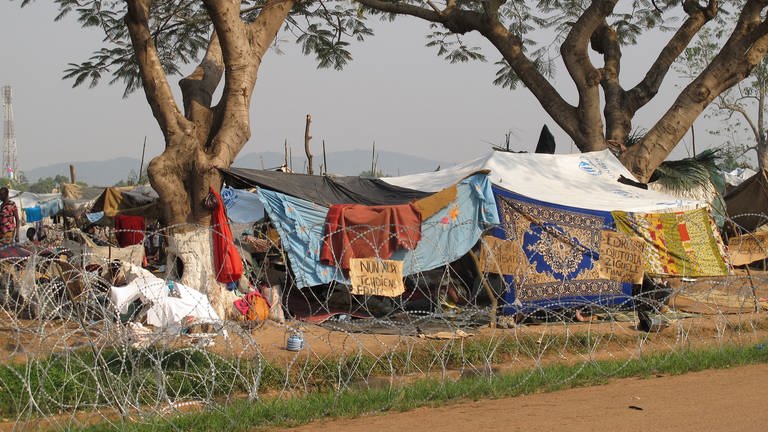 Ein mit Stacheldraht umzäuntes Flüchtlingslager am Rand einer Straße. Als Wände und Dächer dienen Tücher und Planen vom UNHCR