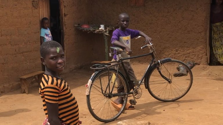 Drei Jungen mit einem Fahrrad vor einem Lehmbau in der Flüchtlingssiedlung
