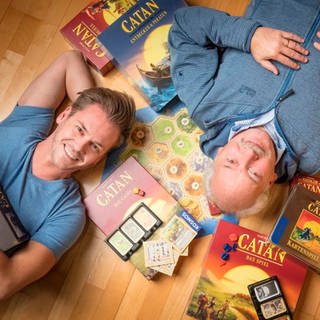Benjamin (li.) und Klaus Teuber (re.), Spieleentwickler, diagonal zueinander auf dem Boden liegend, umgeben von Spielekartons mit der Aufschrift "Catan"