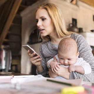 Eine Frau sitzt mit Baby im Arm und Smartphone in der Hand vor einem Notebook
