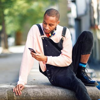 Unsere SWR2 Netztipps für den Sommer 2021: ein junger, schwarzer Mann sitzt mit aufgestütztem Bein auf einer Mauer, an einem sonnigen Tag. Im Hintergrund ist ein Platz in einer Stadt mit Bäumen gesäumt zu sehen. Der junge Mann trägt einen hellrosa Pullover und eine schwarze 78 Latzhose. Er hat einen Kopfhörer um den Hals gehängt und scrollt in seinem Smartphone.