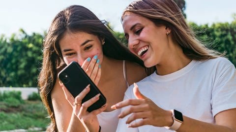 Zwei weiße Teenie-Mädchen mit längeren Haaren blicken auf ein Smartphone und amüsieren sich.