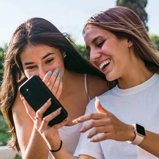 Zwei weiße Teenie-Mädchen mit längeren Haaren blicken auf ein Smartphone und amüsieren sich.