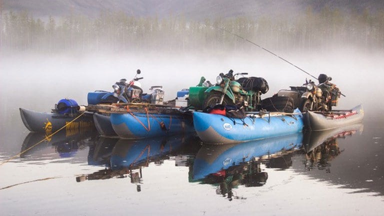 Motorräder stehen auf einem großen Floß, das im Wasser liegt. Der Hintergrund ist neblig