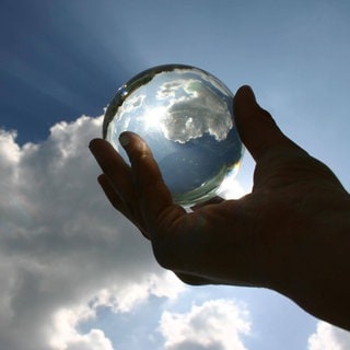 Eine Hand hält eine Glaskugel gegen einen blauen Himmel mit weißen Wolken