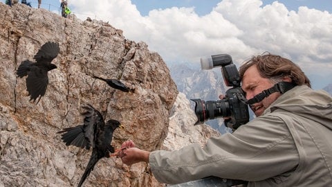 Das Foto zeigt Hans-Jürgen Burkard vor dem Gipfel der Zugspitze. Er hat Alpendohlen mit Rosinen in der Hand angelockt, um sie mit dem Weitwinkel vor dem Gipfel zu fotografieren. 