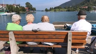 Rentner sitzen an einem sonnigen Tag auf einer Bank und geniessen den Blick auf den Tegernsee in Rottach Egern.