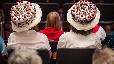 Zwei Frauen tragen Hüte in Form von Schwarzwälder Kirschtorten bei der Abschlussfeier des 121. Deutschen Wandertags in der Schwabenlandhalle.