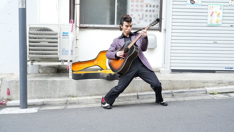 Ein Mann spielt Gitarre auf einer Straße  Ausschnitt aus dem japanischen Film: LONESOME VACATION von Atsuro SHIMOYASHIRO. 