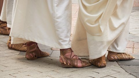 Fuße dreier Personen in weißer, bodenlanger Toga. Sie tragen römische Sandalen, sogenannte caligae.
