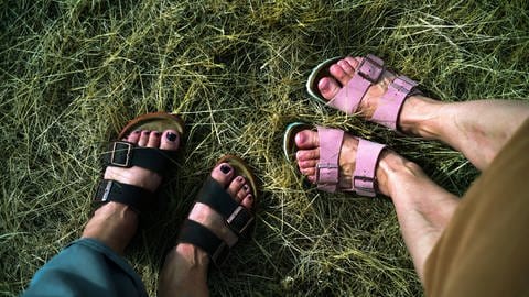 Im Gras Füße von zwei weiblich gelesenen Personen in Sandalen nach Art der Marke Birkenstock. Links eine Frau mit dunklen Schuhen und dunkel lackierten Fußnägeln, links eine Frau in hellrosa Sandalen