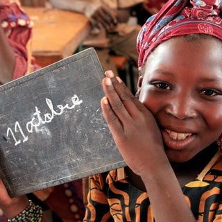 Ein Mädchen in Niger hält eine kleine Tafel, auf der «11. Oktober» geschrieben steht, in ihrem Klassenzimmer hoch