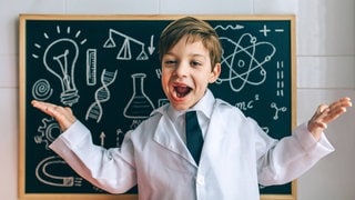 fröhliches KInd, bekleidet mit einem Lehrerkittel, vor einer Schultafel