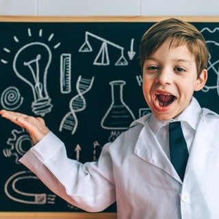 fröhliches KInd, bekleidet mit einem Lehrerkittel, vor einer Schultafel