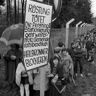 Demonstranten mit einem "Rüstung tötet"-Plakat vor dem Sicherheitszaun des Militärgeländes.