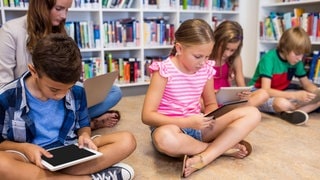 Grundschulkinder sitzen auf dem Boden und schauen auf Tablets