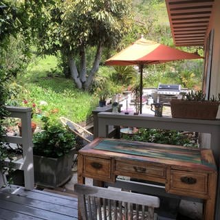 Kleiner Holz-Schreibtisch auf der Terrasse, Blick in den Garten mit alten Bäumen, Blumen und auf einen orange-farbenen Sonnernschirm